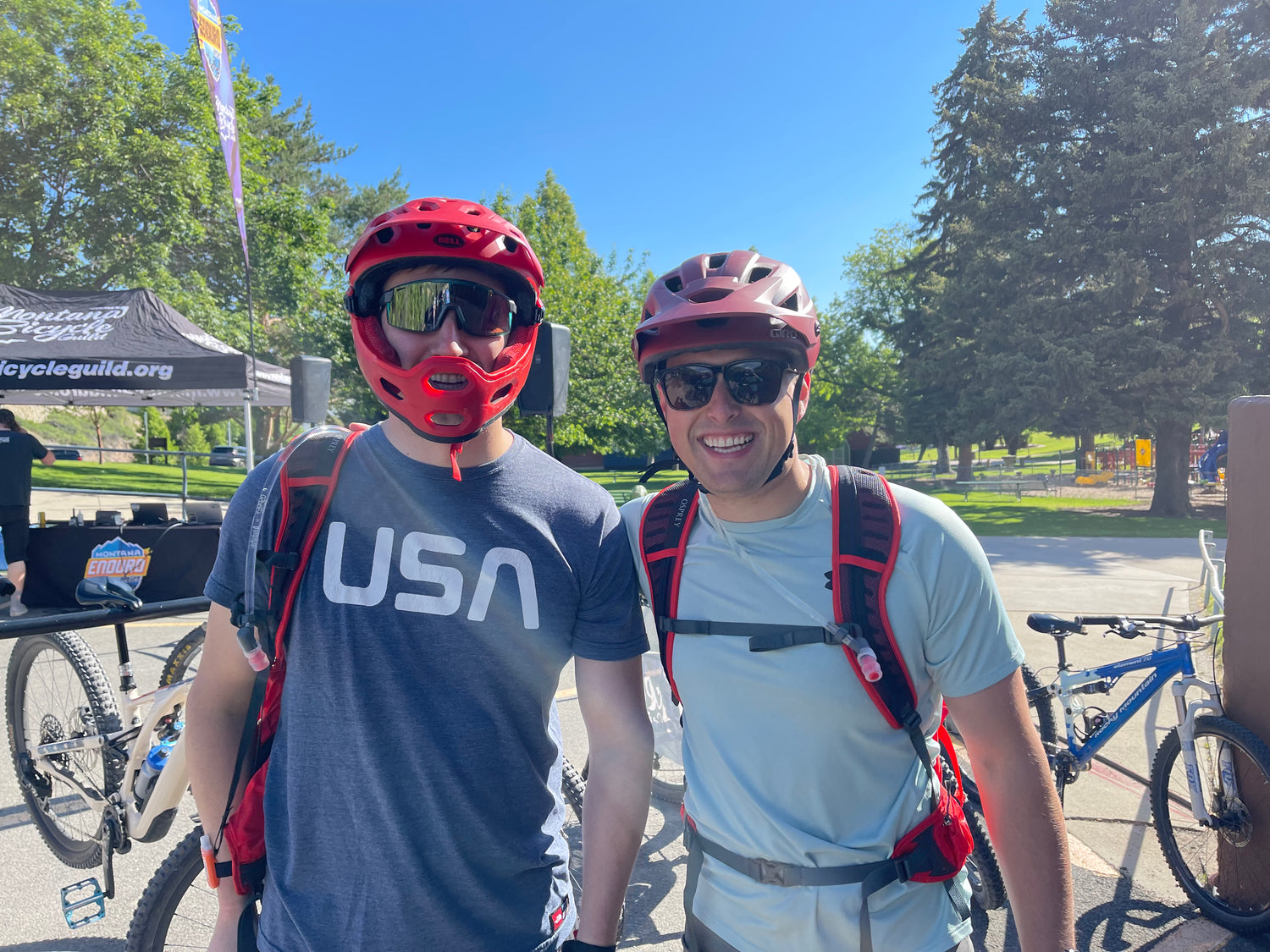 Two men wearing red helmets and sunglasses standing next to bicycles in a park setting.